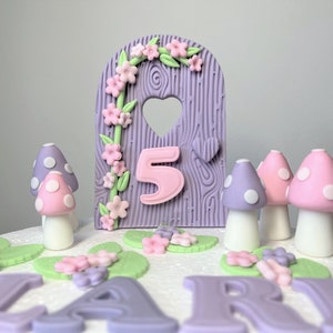 May include: Purple and pink cake topper featuring a wooden door, flowers, and the number 5. There are also purple and pink mushrooms with white polka dots.