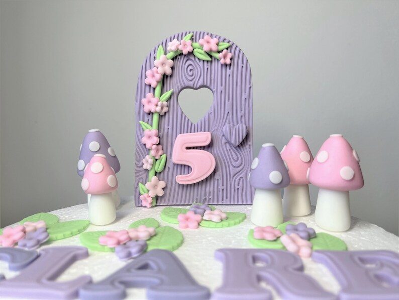 May include: Purple and pink cake topper featuring a wooden door, flowers, and the number 5. There are also purple and pink mushrooms with white polka dots.