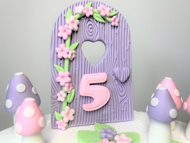 May include: Purple and pink fairy door cake topper with a heart-shaped window and a pink number 5. The door is decorated with a green vine and pink flowers. There are also purple and pink mushrooms with white polka dots.