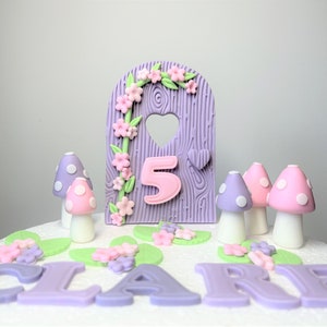 May include: Purple and pink fairy door cake topper with a number 5 and the name "CLAIRE" written in icing. The topper is surrounded by pink and green flowers and mushrooms.