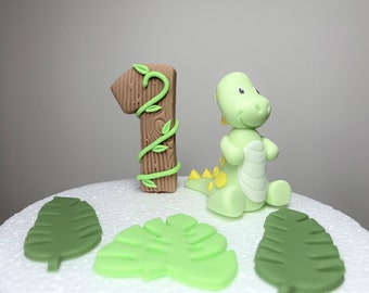 May include: A green dinosaur cake topper with a yellow belly and a brown number one cake topper with green vines. The cake topper is on a white cake board with green leaves.