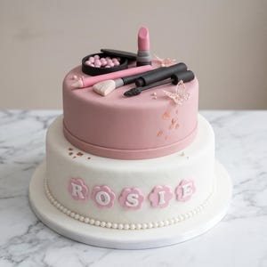 Könnte beinhalten: Eine zweistöckige Torte, die mit Elementen zum Thema Make-up verziert ist. Die obere Etage ist rosa und zeigt einen Lippenstift, Mascara, einen Make-up-Pinsel und ein Etui mit rosa Perlen. Die untere Etage ist weiß mit dem Namen "Rosie" in rosa blütenförmigen Buchstaben.