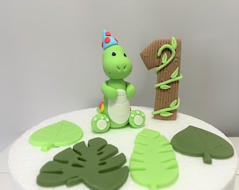 May include: Green dinosaur cake topper wearing a blue and red party hat, with a brown number one cake topper decorated with green vines, and green leaves.