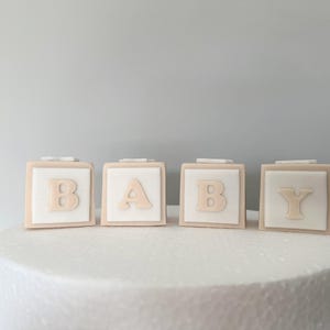 May include: Four square blocks spelling "BABY" in light beige letters. Each block has a white center with a light beige border. The blocks are on a white, round surface, likely a cake.