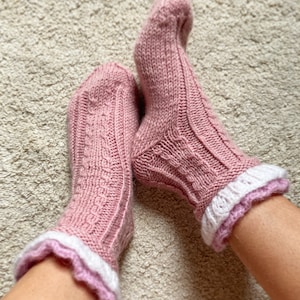 May include: A pair of pink knitted socks with white and pink ruffled trim. The socks are worn on a person's feet.