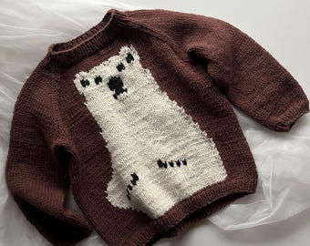 Puede incluir: Un suéter de punto marrón con un diseño de oso polar blanco. El suéter tiene mangas largas y cuello redondo.