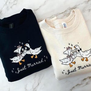 May include: Two sweatshirts, one black and one cream, each with an embroidered design of geese celebrating. The geese are holding champagne glasses and wearing bows. The words "Just Married" are embroidered below the geese.