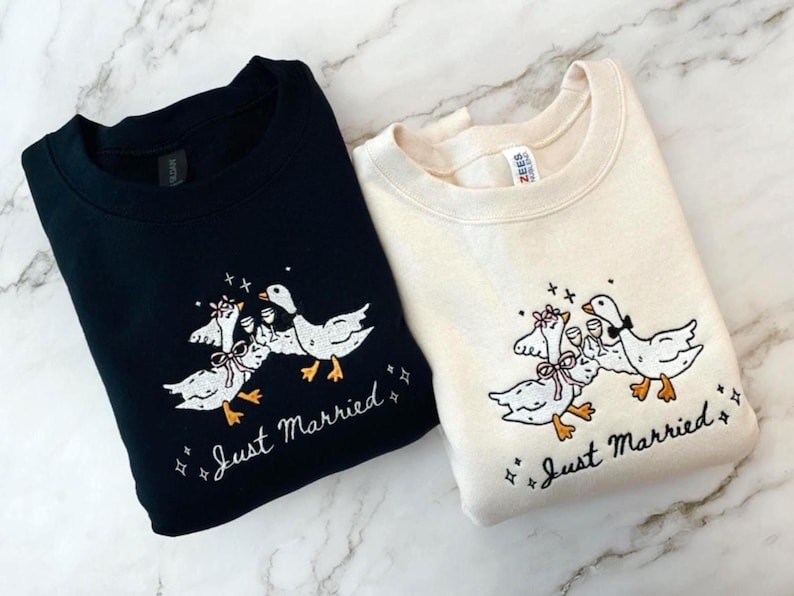 May include: Two sweatshirts, one black and one cream, each with an embroidered design of geese celebrating. The geese are holding champagne glasses and wearing bows. The words "Just Married" are embroidered below the geese.
