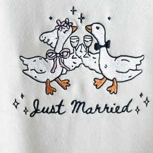 May include: Cream-colored sweatshirt with an embroidered design of two geese toasting with wine glasses. The geese are adorned with bows and a flower crown. The words "Just Married" are embroidered below the geese.
