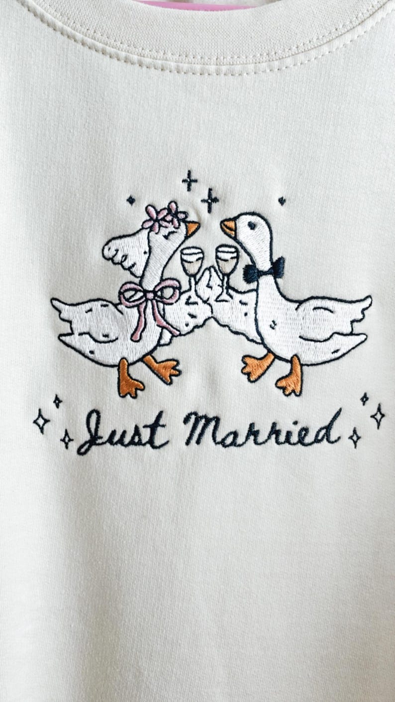 May include: Cream-colored sweatshirt with an embroidered design of two geese toasting with wine glasses. The geese are adorned with bows and a flower crown. The words "Just Married" are embroidered below the geese.