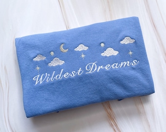 Op de afbeelding: Een gevouwen blauw stoffen item met geborduurde hemelse ontwerpen, waaronder wolken, sterren en een halve maan. De woorden "Wildest Dreams" zijn in cursief geborduurd. Het item heeft een lichtblauwe kleur.