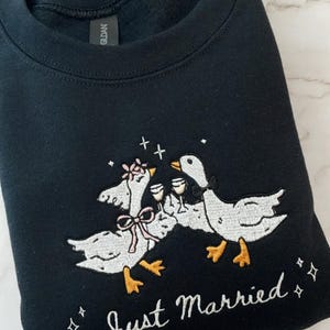 May include: Black sweatshirt with an embroidered design of two geese toasting with champagne glasses. The geese are wearing bows and flowers, with the words "Just Married" in script below. A fun, celebratory design.