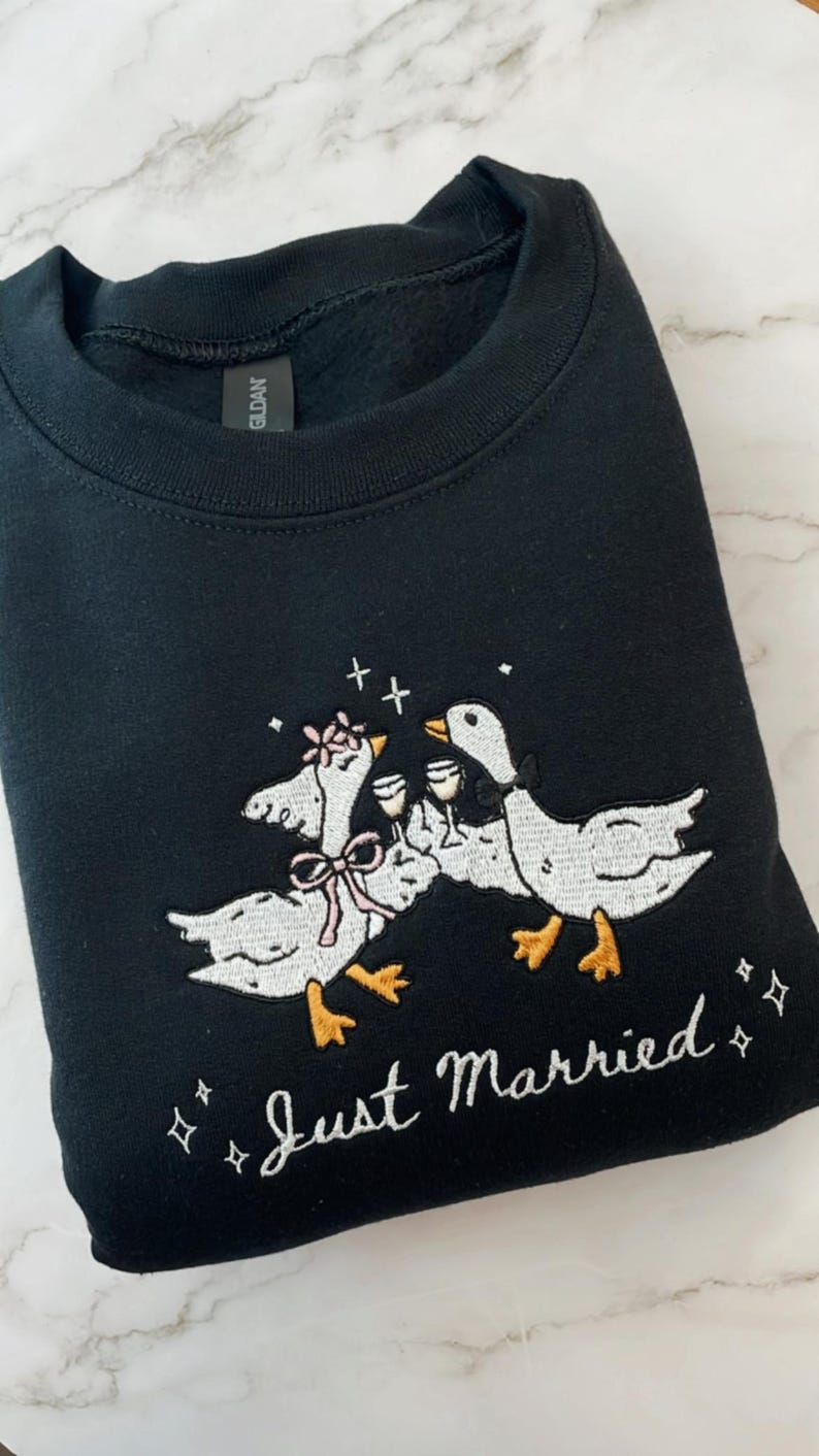 May include: Black sweatshirt with an embroidered design of two geese toasting with champagne glasses. The geese are wearing bows and flowers, with the words "Just Married" in script below. A fun, celebratory design.