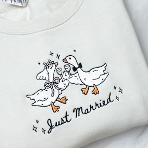 May include: Cream-colored sweatshirt with an embroidered design of three geese toasting with wine glasses. The geese are adorned with bows and a bow tie. The words "Just Married" are embroidered below the geese, with star accents.