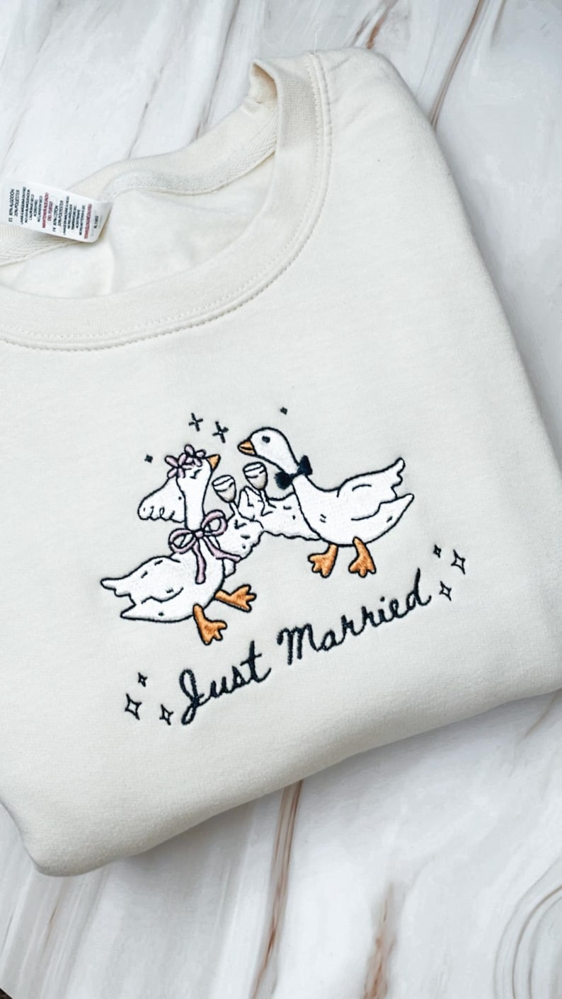 May include: Cream-colored sweatshirt with an embroidered design of three geese toasting with wine glasses. The geese are adorned with bows and a bow tie. The words "Just Married" are embroidered below the geese, with star accents.