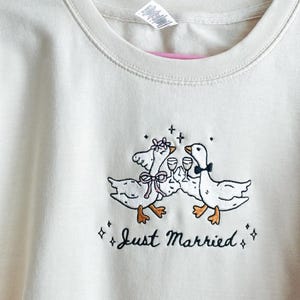 May include: Cream-colored sweatshirt with an embroidered design of two geese toasting with wine glasses. The geese are adorned with bows and floral accents. The text "Just Married" is embroidered below the geese.