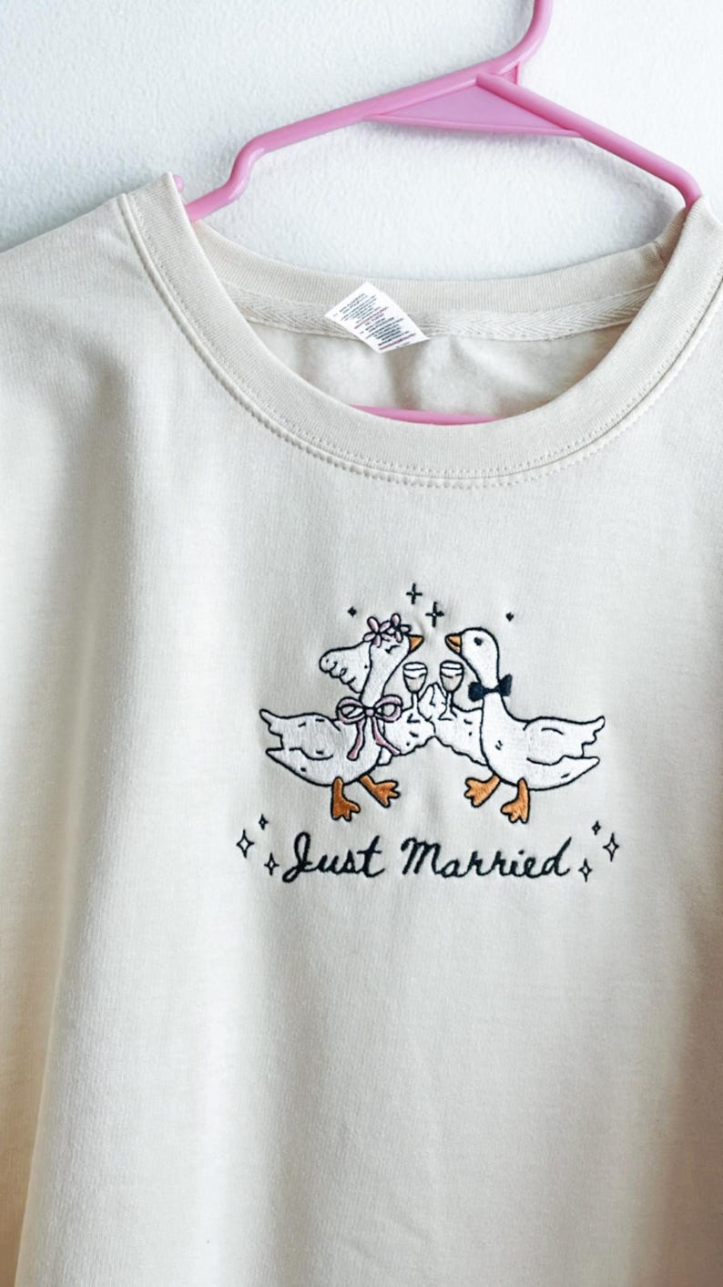 May include: Cream-colored sweatshirt with an embroidered design of two geese toasting with wine glasses. The geese are adorned with bows and floral accents. The text "Just Married" is embroidered below the geese.