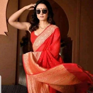 May include: A woman wearing a vibrant red sari with intricate gold embroidery. The sari drapes elegantly, showcasing the detailed patterns. She wears sunglasses and a delicate necklace, posing in a well-lit setting.