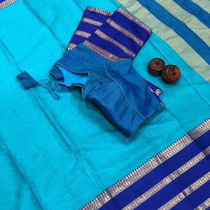 May include: A turquoise blue saree with a blue and gold border. The saree is folded and laid out on a dark surface. A matching blue blouse is also included.