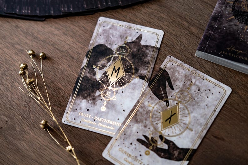 Puede incluir: Dos cartas de tarot con detalles dorados y dise&ntilde;os de acuarela en blanco y negro. La carta de la izquierda tiene la letra "M" en un diamante dorado y el texto "TRUST - PARTNERSHIP". La carta de la derecha tiene la letra "X" en un diamante dorado y el texto "GEBO".