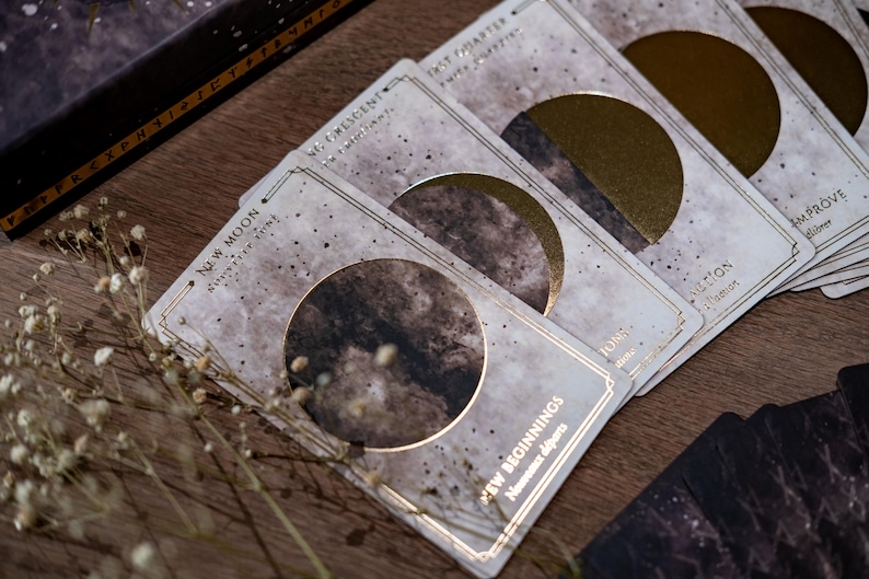 Puede incluir: Un juego de cartas con un tema celestial. Cada carta presenta una fase diferente de la luna con un dise&ntilde;o de l&aacute;mina dorada. Las cartas est&aacute;n dispuestas sobre una superficie de madera con un peque&ntilde;o ramo de flores blancas.