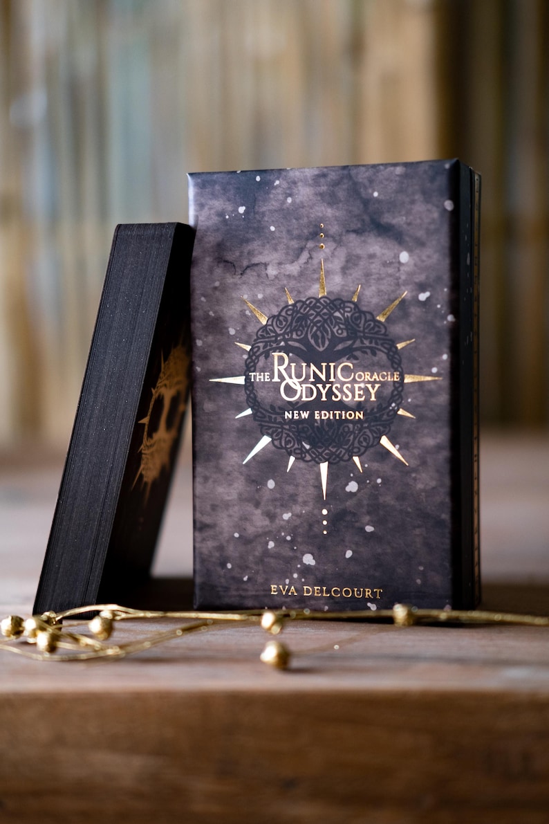 Puede incluir: Una caja negra con letras doradas que dicen "The Runic Oracle Odyssey New Edition" de Eva Delcourt. La caja tiene un fondo gris oscuro con detalles dorados.