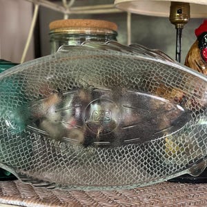 Op de afbeelding: Een doorzichtige glazen serveerschaal in de vorm van een vis, voorzien van een gestructureerd schubbenpatroon over het lichaam, een gedefinieerde staartvin en een centraal rond embleem met reliëftekst. De schaal heeft een gladde rand en een holle vorm.