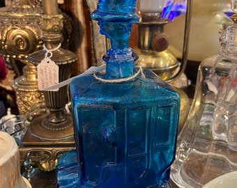 Peut inclure: Une carafe en verre bleu cobalt en forme de diligence ancienne avec un haut-de-forme servant de bouchon. La bouteille présente des détails moulés incluant des roues, des fenêtres et une porte, avec une ficelle nouée autour du goulot.