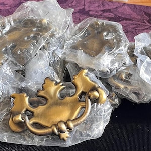 Puede incluir: Una colección de tiradores de cajón de metal en tono latón, cada uno con una placa decorativa ornamentada con un asa basculante. Varias de estas piezas de herraje están envueltas individualmente en bolsas de plástico transparente.
