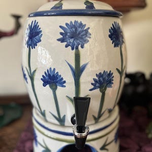 Pode incluir: Um dispensador de bebidas de cerâmica com esmalte cor de creme e motivos de centáureas azuis pintados à mão. O recipiente cilíndrico possui uma tampa a condizer, uma torneira de plástico preta na frente e uma base robusta com detalhes em azul e verde.