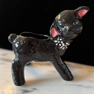 May include: A glossy black ceramic planter shaped like a lamb with a textured wool body. The lamb features pink-painted inner ears and a white-painted collar with small dots around its neck, with an opening on its back for small plants.