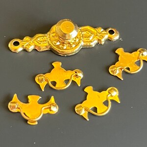 May include: A set of five gold-colored metal furniture hardware pieces. One piece is a long, ornate drawer pull with a central knob and two screw holes. The other four pieces are smaller, matching drawer pulls with curved, swinging handles.