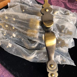 May include: A collection of antique brass-toned metal drawer pulls, each featuring an elongated, curved handle with decorative, scalloped ends and small circular accents. One pull is unwrapped, while the others remain in clear plastic packaging.