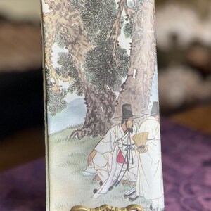May include: A rectangular fabric panel featuring a printed illustration of two figures in traditional white robes and black hats standing beneath a large tree. One figure holds a small yellow fan. The panel is displayed on a gold-toned metal stand.