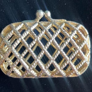 Puede incluir: Un broche de metal dorado con forma de pequeño bolso de mano rectangular. La superficie presenta un patrón de celosía o cuerda cruzada en relieve por todo el cuerpo, rematado con un mecanismo de cierre de doble botón en la parte superior.