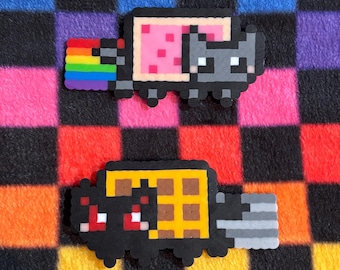 May include: Two pixel art figures made of plastic beads. The top figure is a gray cat with a pink rectangular body and a rainbow trail. The bottom figure is a black cat with a yellow waffle body and a gray trail, featuring red eyes.
