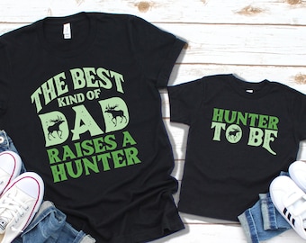 May include: Two black t-shirts with green text and a moose graphic. The larger shirt says "The Best Kind of Dad Raises a Hunter". The smaller shirt says "Hunter to Be".