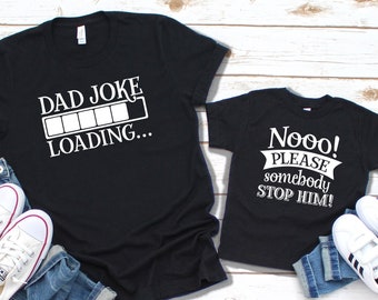 May include: Two black t-shirts with white text. One says "DAD JOKE LOADING..." and the other says "Nooo! PLEASE somebody STOP HIM!". The shirts are paired with blue jeans and white sneakers.