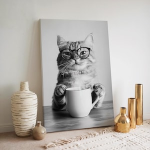 Op de afbeelding: Zwart-witfoto van een kat met een bril die een witte mok vasthoudt. De kat zit op een grijs oppervlak.