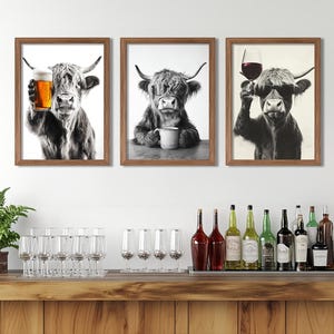 May include: Three black and white framed prints of a Highland cow with different beverages. The cow in the left print is holding a glass of beer, the cow in the centre print is holding a mug of coffee, and the cow in the right print is holding a glass of wine.