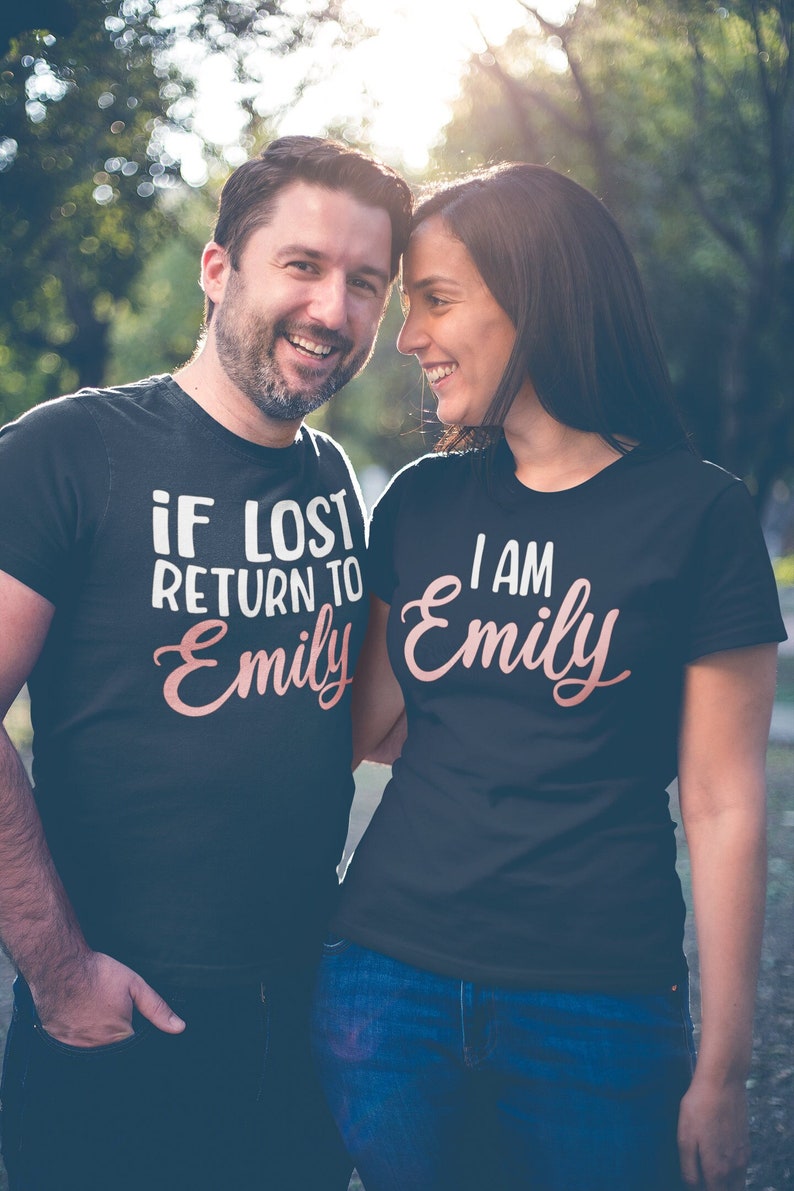 May include: A couple wearing matching black t-shirts with a pink design. The man's shirt says "If lost return to Emily" and the woman's shirt says "I am Emily".