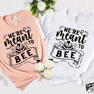 May include: Two t-shirts with a black and white graphic design. The graphic says "We're meant to bee" with a heart and two bees. One shirt is peach and the other is white.