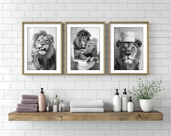 May include: Three black and white framed prints of a lion. The lion in the left print is brushing its teeth. The lion in the center print is sitting on a toilet and reading a newspaper. The lion in the right print is wearing a white towel on its head.