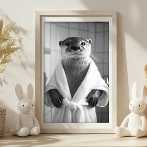 May include: A black and white photo of an otter wearing a white bathrobe. The otter is looking directly at the camera with a serious expression. The photo is framed in a white frame.