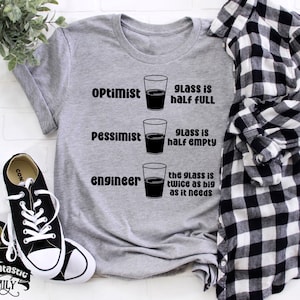May include: Grey t-shirt with a humorous graphic of three glasses of liquid. The text reads "Optimist, glass is half full", "Pessimist, glass is half empty", and "Engineer, the glass is twice as big as it needs".