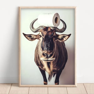 May include: A framed art print featuring a wildebeest with a roll of toilet paper balanced on its horns. The wildebeest has brown and black fur, with curved horns and a serious expression. The print is in a wooden frame.