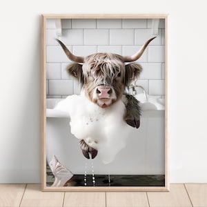 May include: A framed print featuring a Highland cow taking a bubble bath. The cow, with long, shaggy brown fur and large horns, is submerged in a white bathtub filled with foamy bubbles. The background shows white tiles.