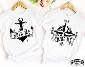 May include: Two white t-shirts with black graphic designs. The first shirt features an anchor with the text "Hold Me". The second shirt features a compass with the text "Guide Me".