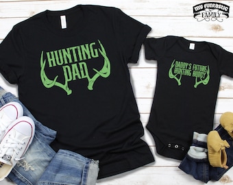 May include: Black t-shirts with green text and antlers. The larger shirt says "Hunting Dad" and the smaller shirt says "Daddy's Future Hunting Buddy".