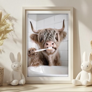 May include: Framed print featuring a Highland cow brushing its teeth in a bathroom setting. The cow has long, shaggy brown fur and large horns. The image is in a white frame, with a neutral color palette. Decorative elements include a bunny figurine and a basket.